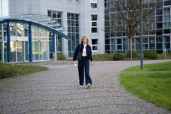 Eine Frau mit blonden, schulterlangen Haaren trägt eine dunkle Kleidung und geht auf einem gepflasterten Weg, umgeben von modernen Gebäuden und grüner Wiese. Die Szene wirkt freundlich und einladend.