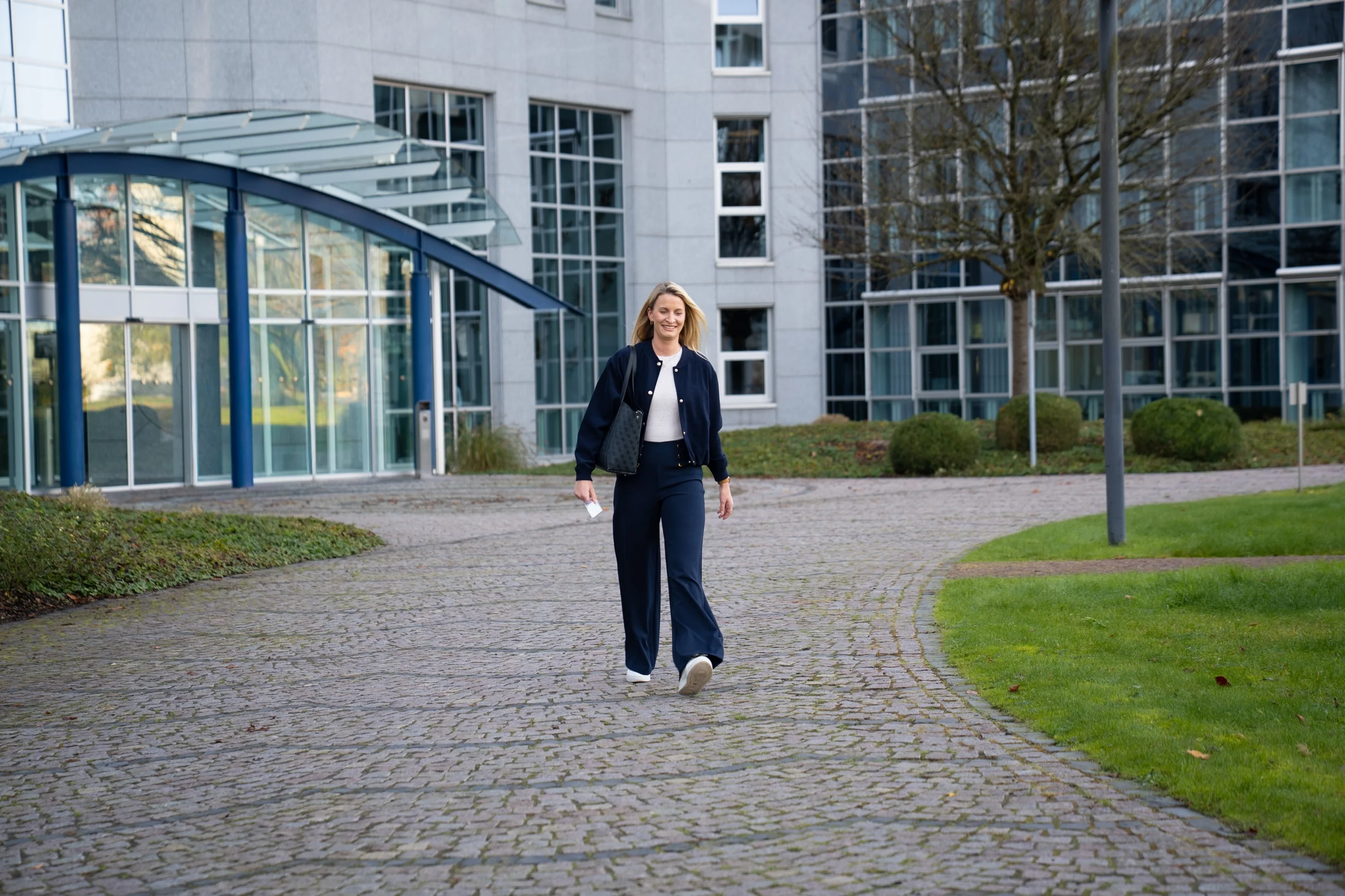 Eine Frau mit blonden, schulterlangen Haaren trägt eine dunkle Kleidung und geht auf einem gepflasterten Weg, umgeben von modernen Gebäuden und grüner Wiese. Die Szene wirkt freundlich und einladend.