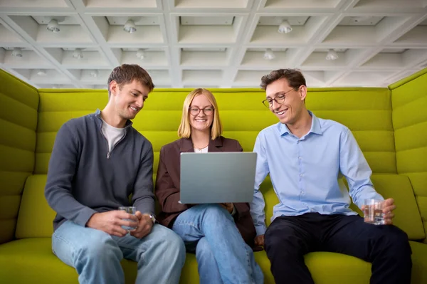 Drei Personen sitzen auf einer grünen Couch. Sie schauen auf einen Laptop und lächeln, während sie sich unterhalten. Jeder hält ein Glas in der Hand, was auf eine entspannte und kollegiale Atmosphäre hinweist.