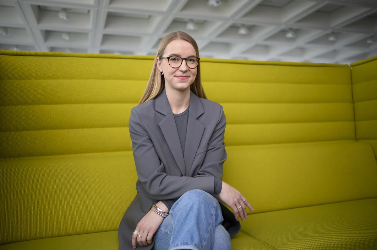 Eine Frau mit langen blonden Haaren sitzt auf einem gelben Sofa. Sie trägt eine Brille, einen grauen Blazer und Jeans. Die Arme sind locker verschränkt. Im Hintergrund ist eine strukturierte, weiße Decke zu sehen.