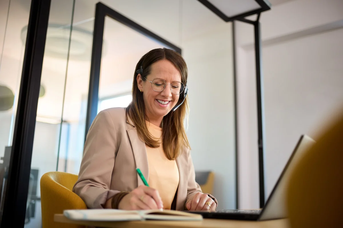Eine lächelnde Frau mit Kopfhörern sitzt an einem Tisch und schreibt in ein Notizbuch, während sie an einem Laptop arbeitet. Sie trägt ein helles Oberteil und einen Blazer. Der Hintergrund zeigt moderne Büroräume mit Glaswänden.
