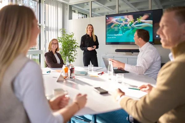In einem modernen Besprechungsraum präsentiert eine Frau vor einem Bildschirm, der die Worte „Firmenkunden“ zeigt. Am Tisch sitzen mehrere Personen, die aufmerksam zuhören. Getränke und Notizen liegen auf dem Tisch. Die Atmosphäre ist professionell und engagiert.