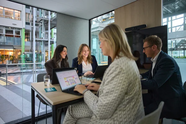 Eine Geschäftssitzung in einem modernen Büroraum. Vier Personen arbeiten an Laptops, während sie miteinander kommunizieren. Der Raum hat große Fenster mit Blick auf eine grüne Umgebung, was eine angenehme und produktive Atmosphäre schafft.