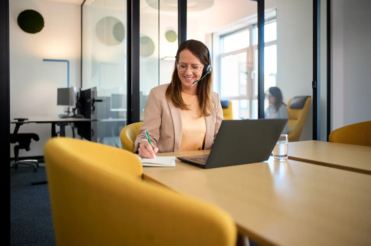 Eine lächelnde Frau sitzt an einem Schreibtisch in einem modernen Büro. Sie trägt ein Headset, arbeitet am Laptop und macht Notizen in einem Notizbuch. Im Hintergrund sind weitere Büroeinrichtungen und eine Kollegin sichtbar.