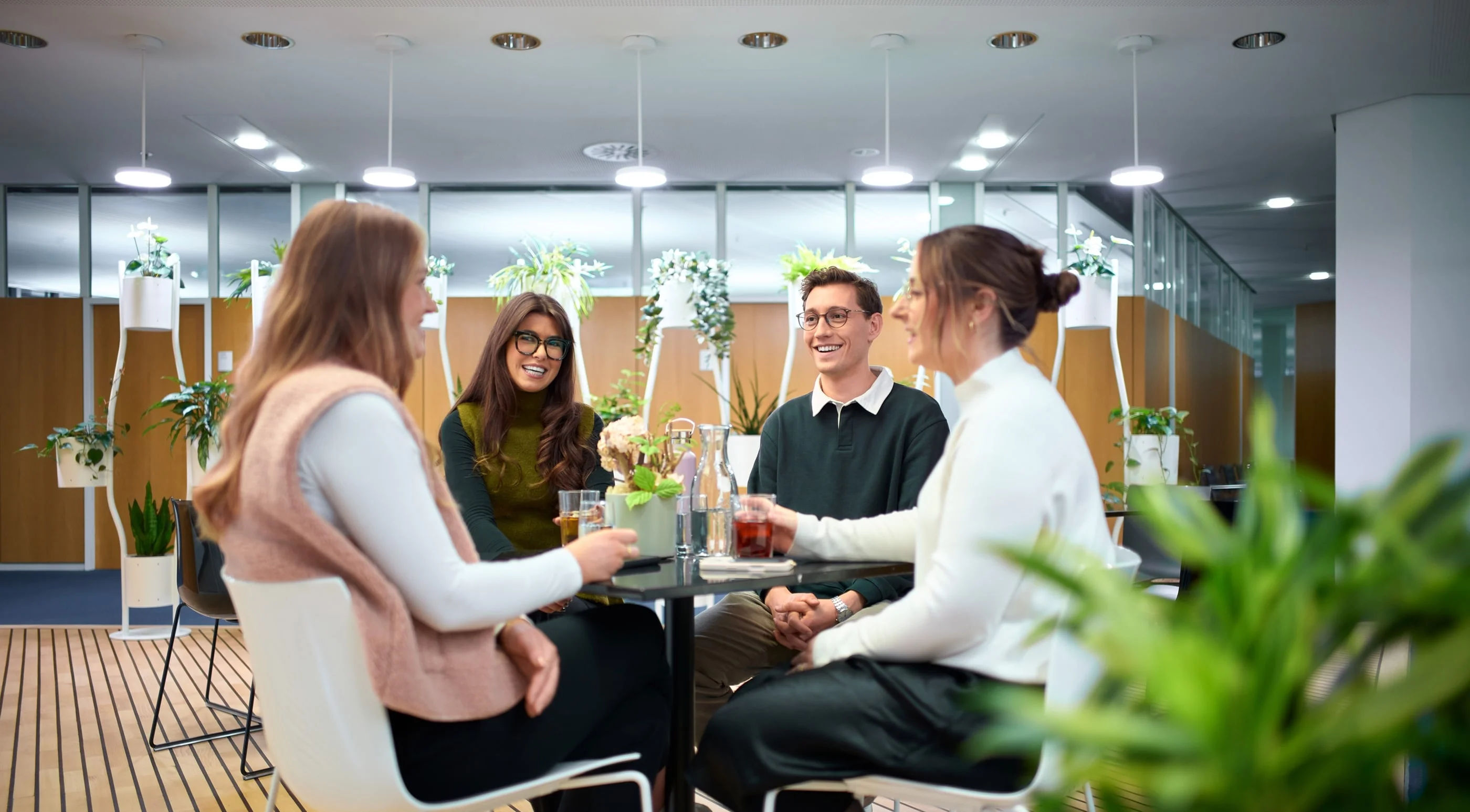Vier Personen sitzen an einem Tisch in einem modernen Büro. Sie lachen und stoßen mit Getränken an. Pflanzen sind im Hintergrund sichtbar, was eine freundliche und entspannte Atmosphäre schafft.
