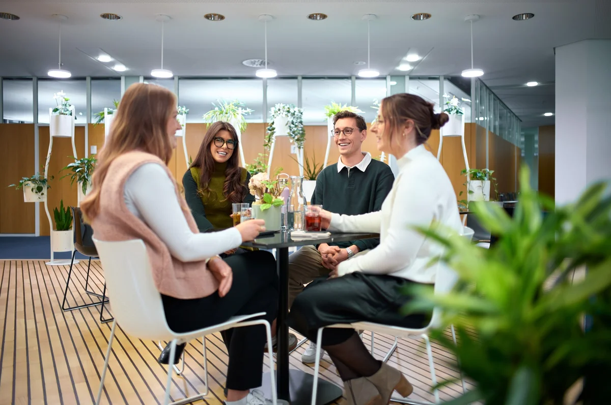 Vier Personen sitzen an einem Tisch in einem modernen Büro. Sie lachen und stoßen mit Getränken an. Pflanzen sind im Hintergrund sichtbar, was eine freundliche und entspannte Atmosphäre schafft.