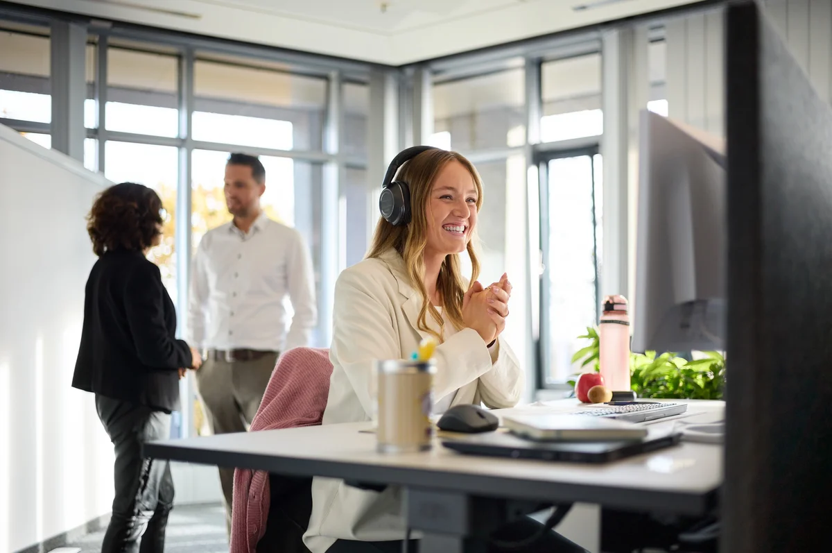 Eine lächelnde Frau trägt Kopfhörer und sitzt an einem Schreibtisch, während sie an einem Computer arbeitet. Im Hintergrund stehen zwei Personen im Gespräch. Pflanzen und Büromaterialien sind auf dem Tisch sichtbar.