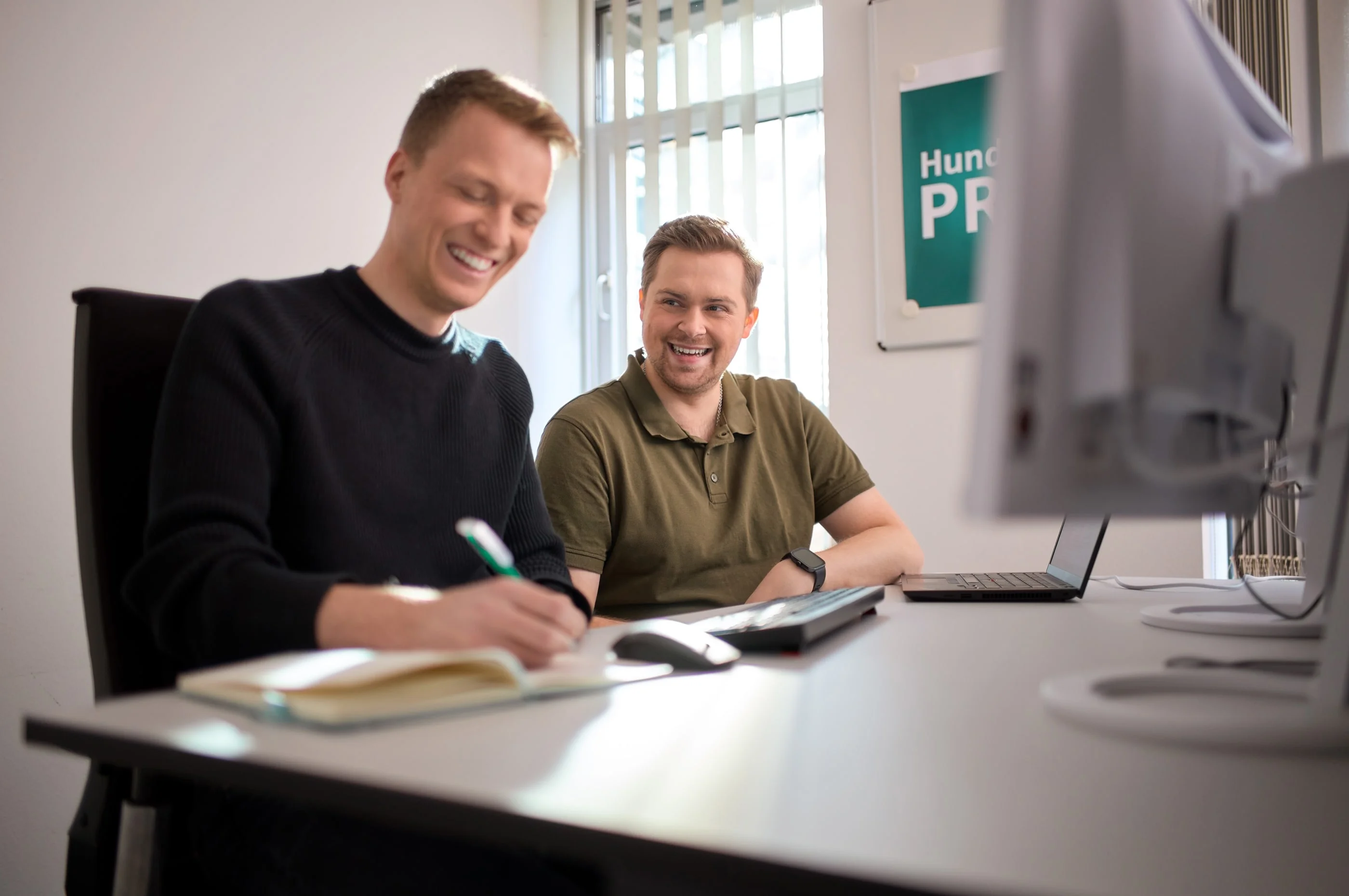 Zwei Männer sitzen an einem Tisch in einem Büro. Der Mann links lächelt und schreibt in ein Notizbuch, während der Mann rechts freundlich schaut. Beide sehen entspannt und engagiert aus, mit einem Computer und einem Notizblock vor ihnen.