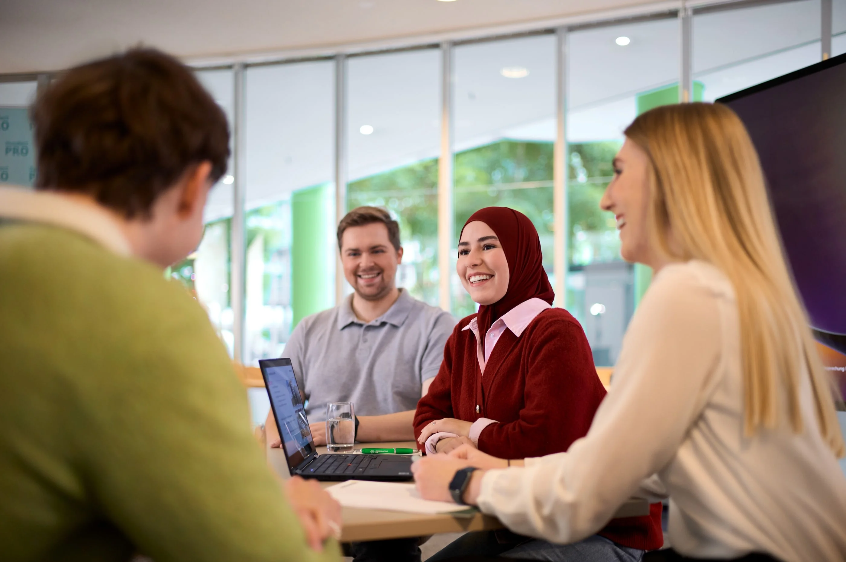 Eine Gruppe von vier Personen sitzt an einem Tisch und diskutiert. Eine Frau mit einem roten Hijab lächelt und spricht, während die anderen interessiert zuhören. Auf dem Tisch steht ein Laptop und zwei Gläser Wasser. Die Atmosphäre wirkt freundlich und kooperativ.