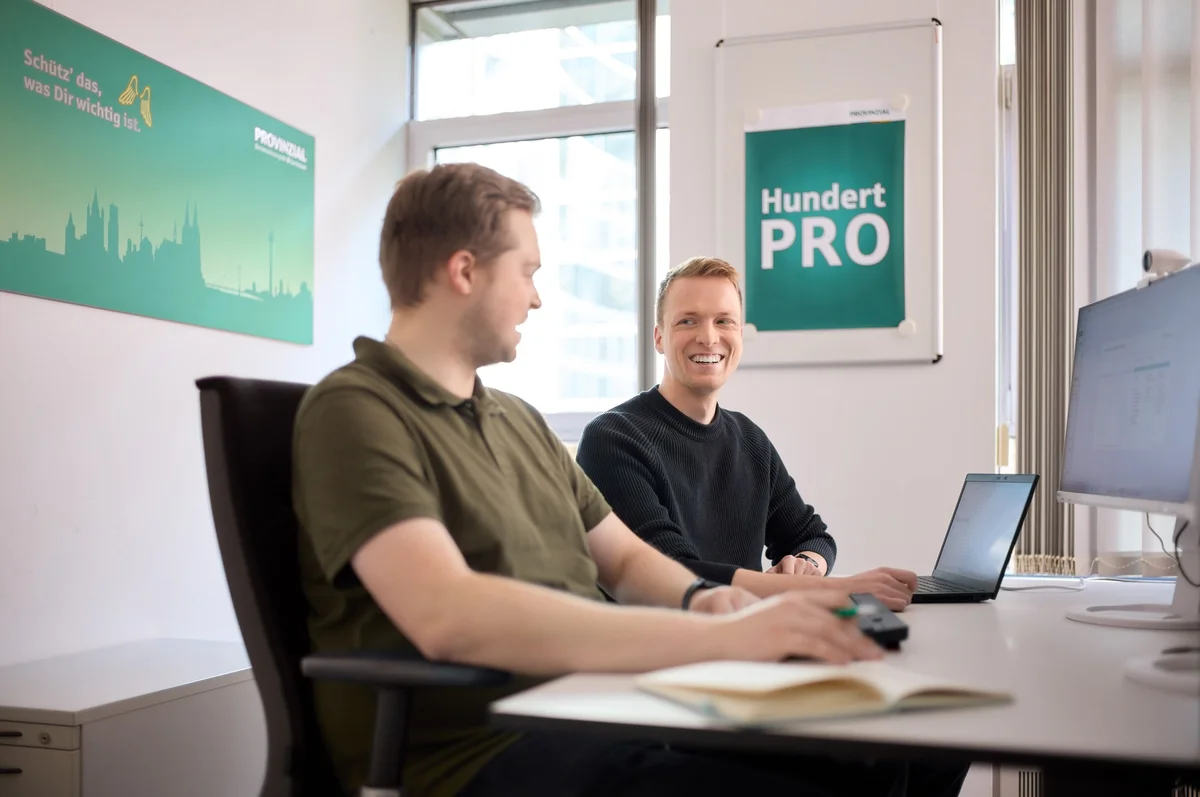 Zwei Männer sitzen an einem Tisch in einem Büro. Einer arbeitet am Laptop, der andere lächelt und schaut ihn an. An der Wand hängen Plakate, darunter eines mit der Aufschrift "Hundert PRO". Der Raum ist hell und modern eingerichtet.