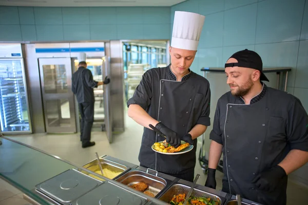 Zwei Köche in einer modernen Küche bereiten Essen vor. Ein Koch, mit einer weißen Kochmütze, serviert auf einem Teller, während der andere Koch aufmerksam zuschaut. Im Hintergrund sind große Kühl- und Kochgeräte sichtbar.