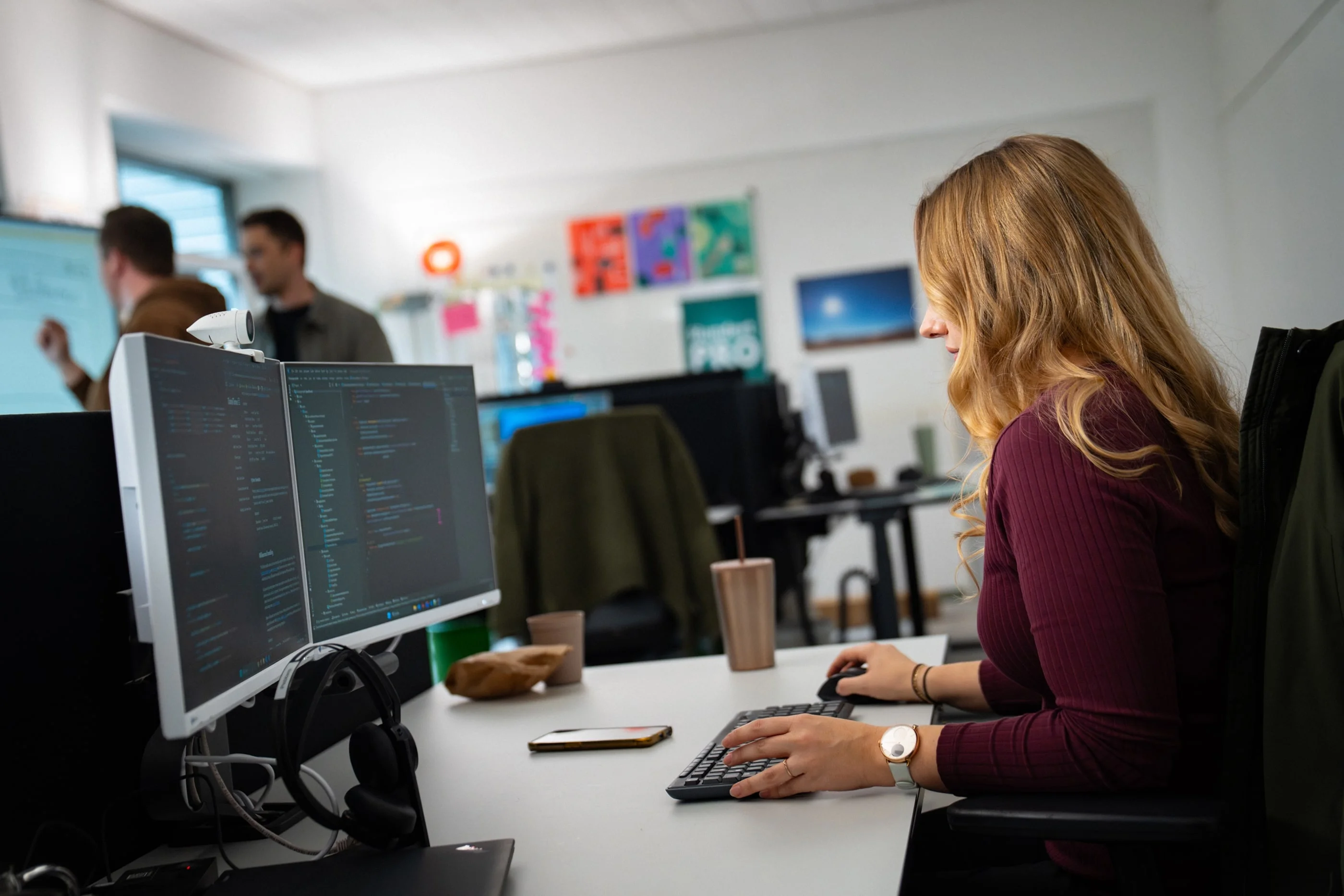 Eine Frau mit langen, blonden Haaren sitzt an einem Schreibtisch und arbeitet am Computer. Auf dem Bildschirm sind Codezeilen sichtbar. Im Hintergrund sind weitere Personen in einem modernen Büro zu sehen. Der Raum ist hell und freundlich eingerichtet.