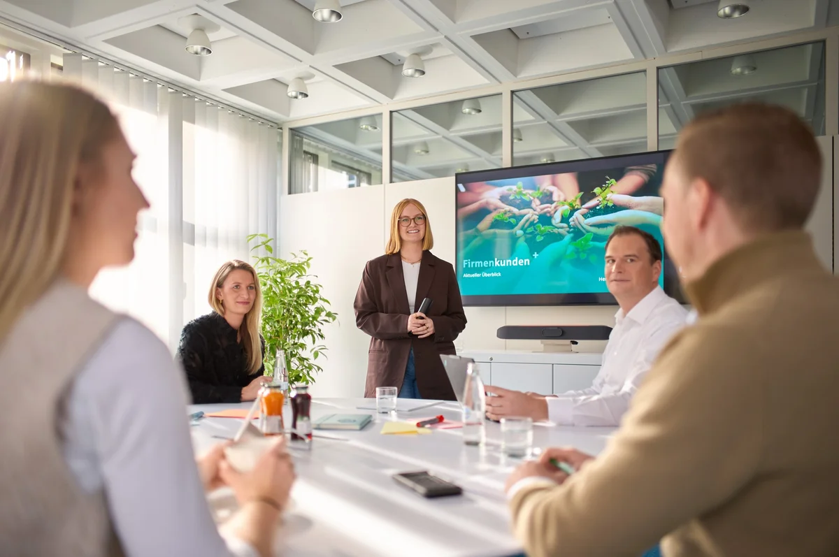 Eine Gruppe von fünf Personen sitzt an einem Tisch in einem modernen Besprechungsraum. Eine Frau steht vorne und präsentiert an einem Bildschirm mit dem Titel „Firmenkunden“. Im Hintergrund sind Pflanzen und Tageslicht zu sehen. Alle Teilnehmer konzentrieren sich auf die Präsentation.