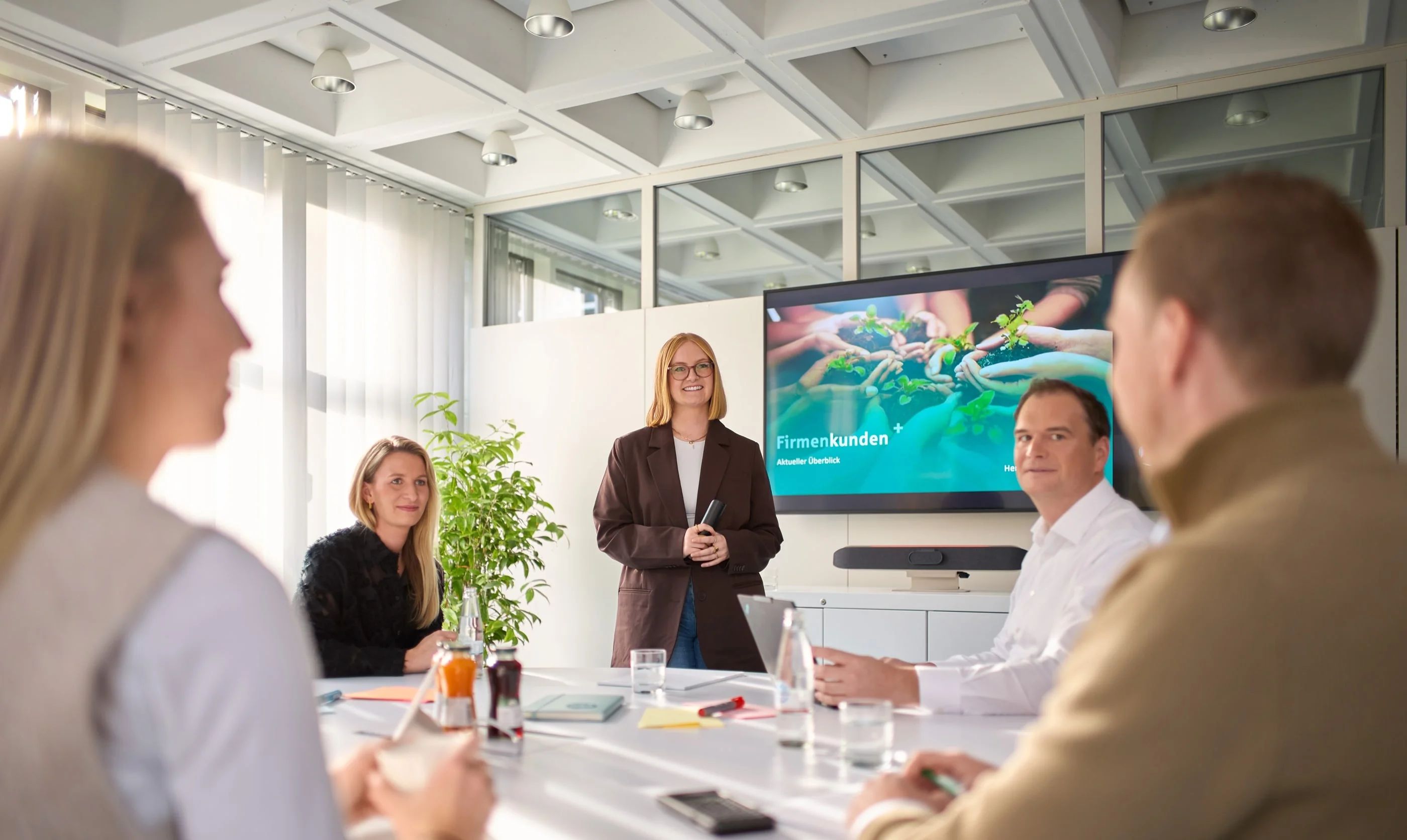Eine Gruppe von fünf Personen sitzt an einem Tisch in einem modernen Besprechungsraum. Eine Frau steht vorne und präsentiert an einem Bildschirm mit dem Titel „Firmenkunden“. Im Hintergrund sind Pflanzen und Tageslicht zu sehen. Alle Teilnehmer konzentrieren sich auf die Präsentation.