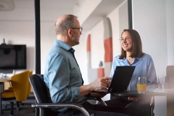Zwei Personen sitzen in einem modernen Besprechungsraum. Ein Mann mit Brille spricht mit einer Frau, die lächelt. Beide haben einen Laptop und ein Glas Orangensaft vor sich. Die Atmosphäre ist freundlich und kollegial.