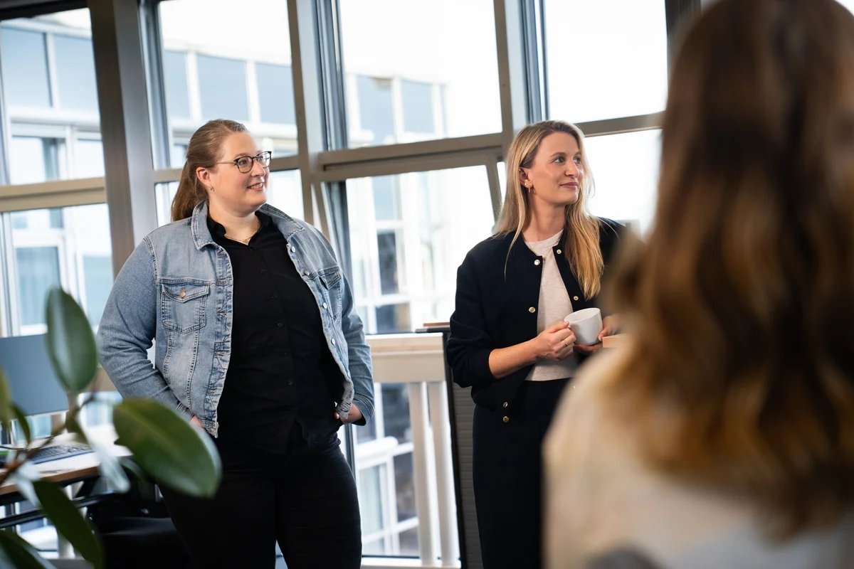 Zwei Frauen stehen in einem modernen Büro. Eine trägt eine Jeansjacke und die andere hält eine Tasse. Sie scheinen an einem Gespräch teilzunehmen, während im Hintergrund Pflanzen und große Fenster sichtbar sind, die Tageslicht hereinlassen.