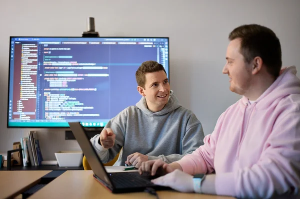 Zwei Menschen sitzen an einem Tisch in einem Büro. Einer von ihnen lächelt und zeigt auf den Laptop, während der andere aufmerksam zuhört. Im Hintergrund ist ein Bildschirm mit Programmiercode sichtbar.