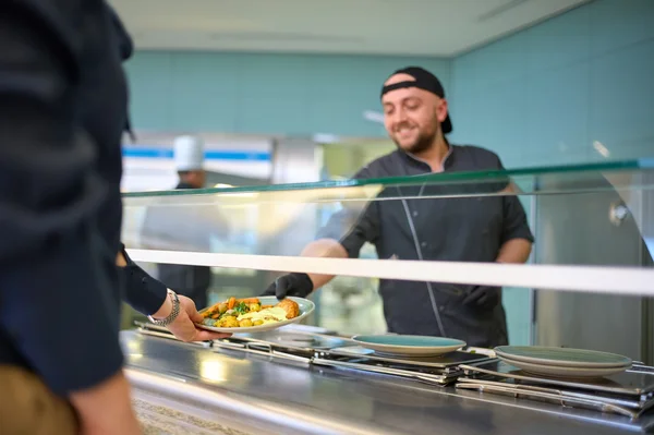 Ein Koch reicht einem Gast ein Gericht über eine Theke. Der Koch lächelt und trägt eine schwarze Kochjacke und Mütze. Der Gast hält einen Teller bereit, um das Essen entgegenzunehmen. Im Hintergrund sind weitere Köche in einer modernen Küche zu sehen.