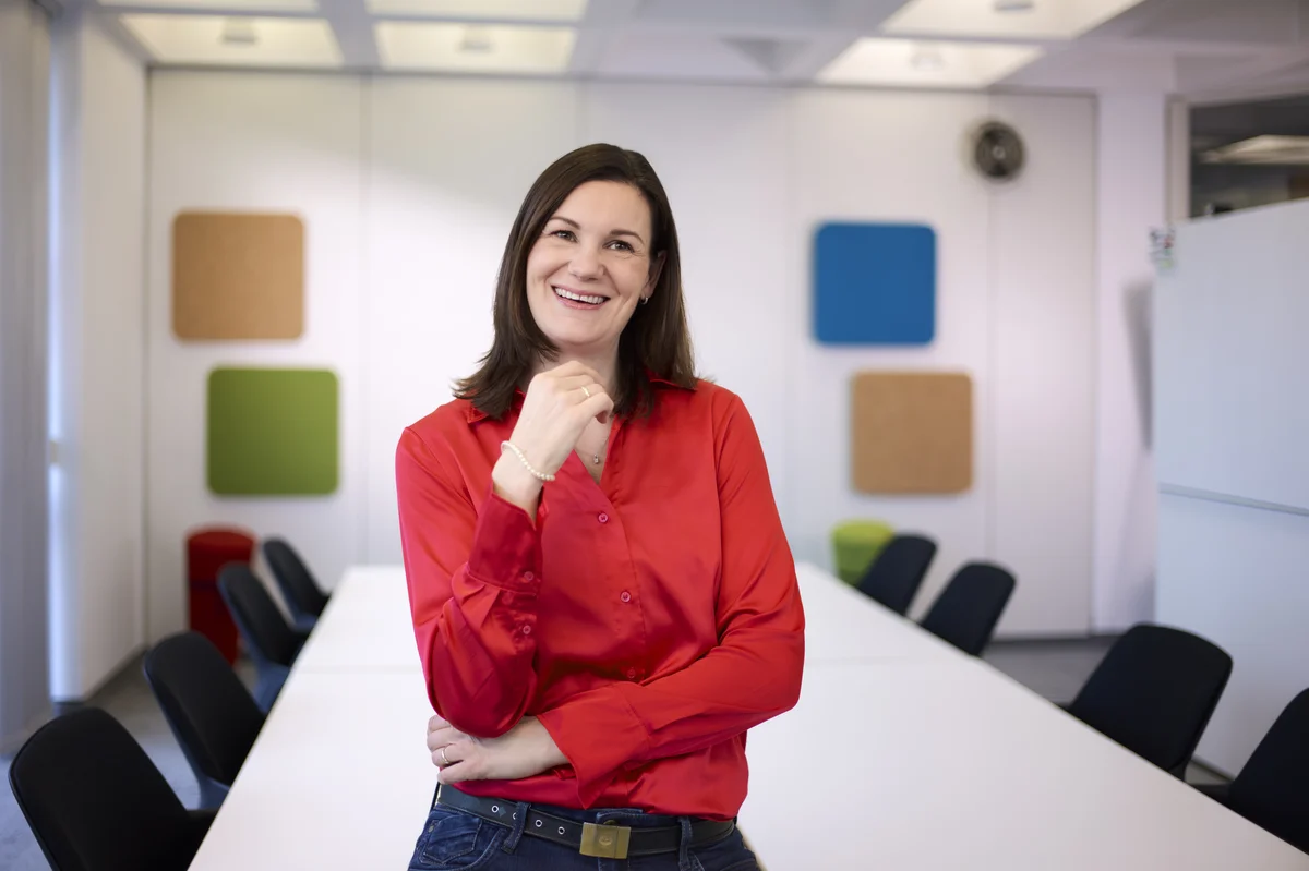 Frau in roter Bluse lächelt in die Kamera, steht in einem modernen Konferenzraum mit großem Tisch und schwarzen Stühlen. An der Wand sind bunte Paneele angebracht.