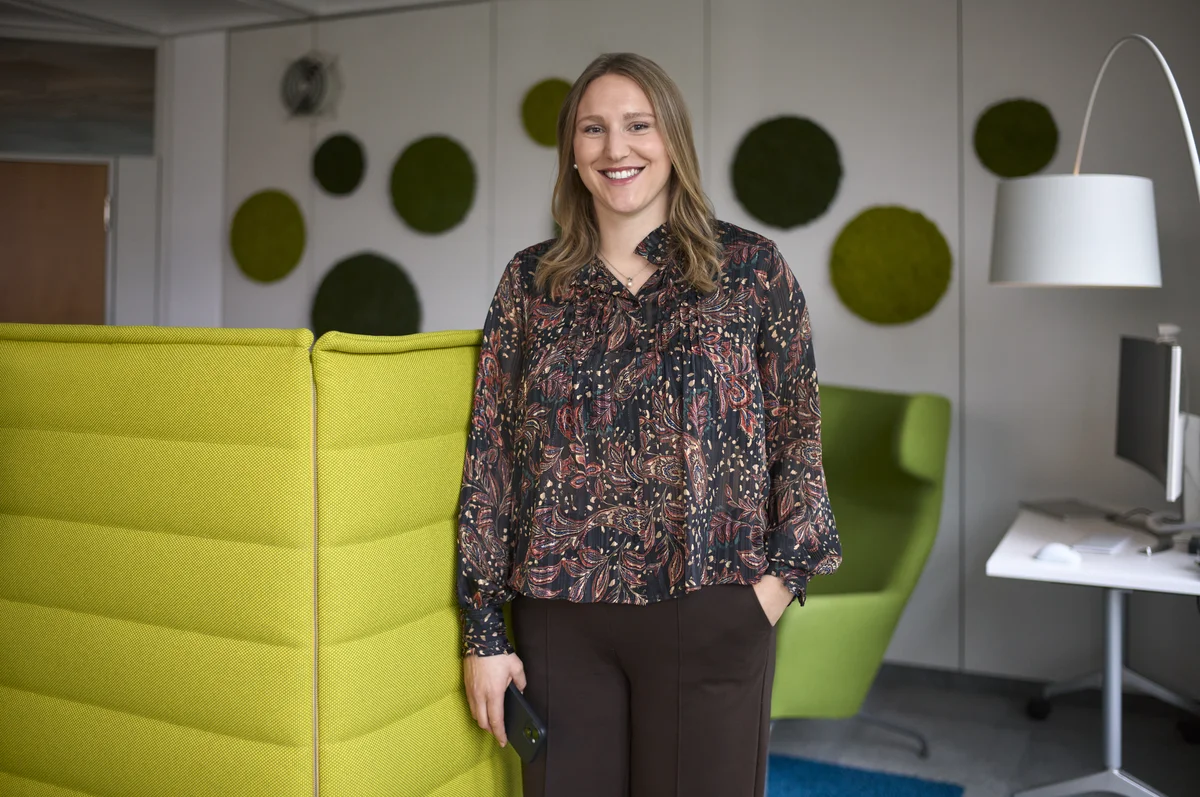 Eine Frau steht lächelnd in einem modernen Büro mit grünen Möbeln und grüner Wanddekoration. Im Hintergrund sind ein Tisch, ein Stuhl und ein Computerbildschirm zu sehen. Sie trägt eine gemusterte Bluse und dunkle Hosen.