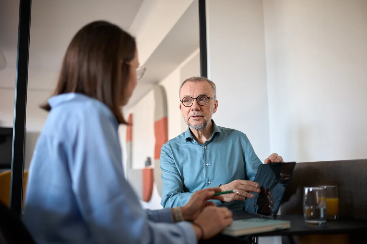 Ein älterer Mann und eine Frau sitzen an einem Tisch. Der Mann hält ein Dokument in der Hand und spricht offenbar über dessen Inhalte, während die Frau ihm aufmerksam zuhört. Auf dem Tisch stehen ein Glas Saft und ein Notizbuch.