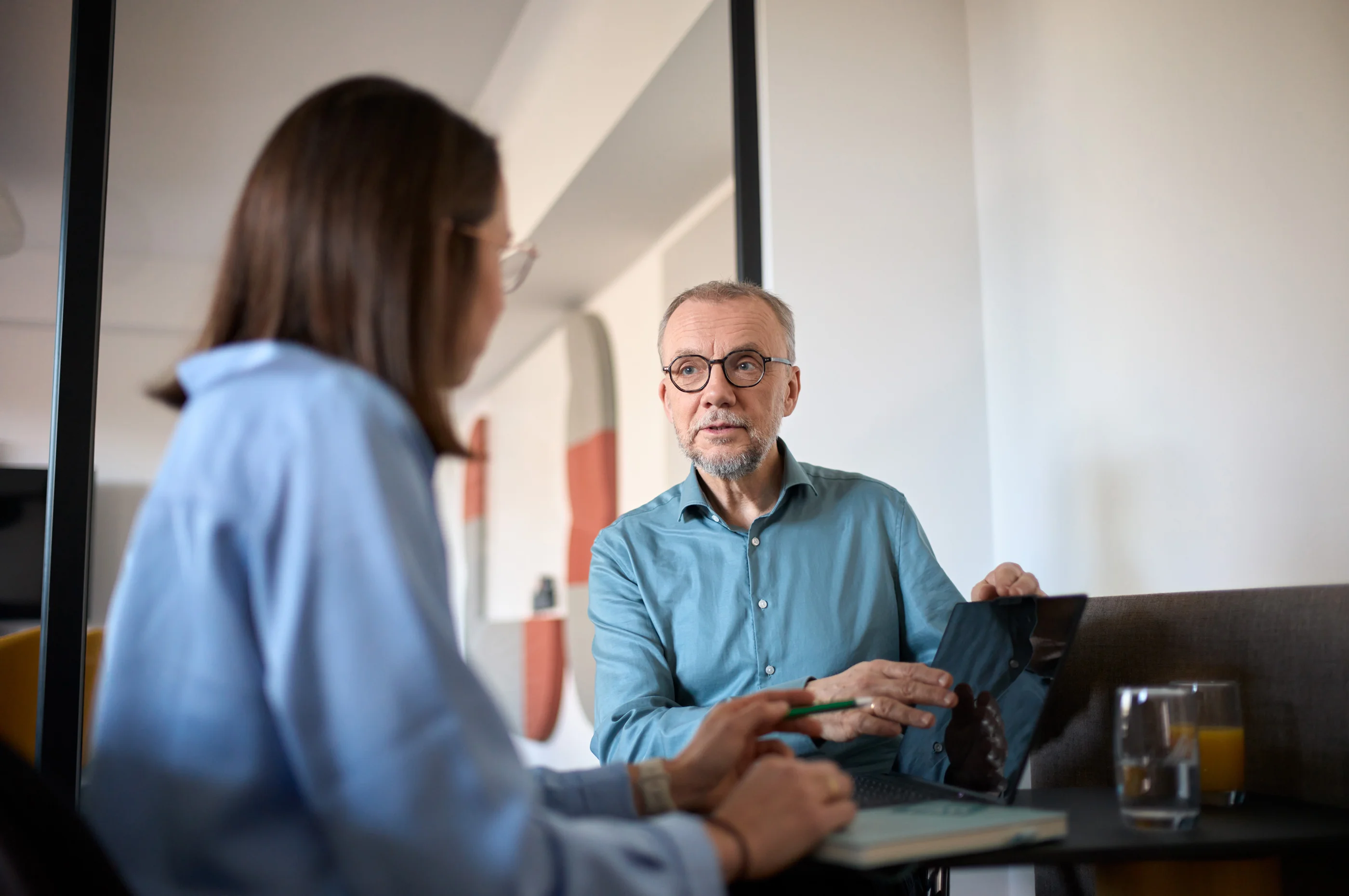 Ein älterer Mann und eine Frau sitzen an einem Tisch. Der Mann hält ein Dokument in der Hand und spricht offenbar über dessen Inhalte, während die Frau ihm aufmerksam zuhört. Auf dem Tisch stehen ein Glas Saft und ein Notizbuch.