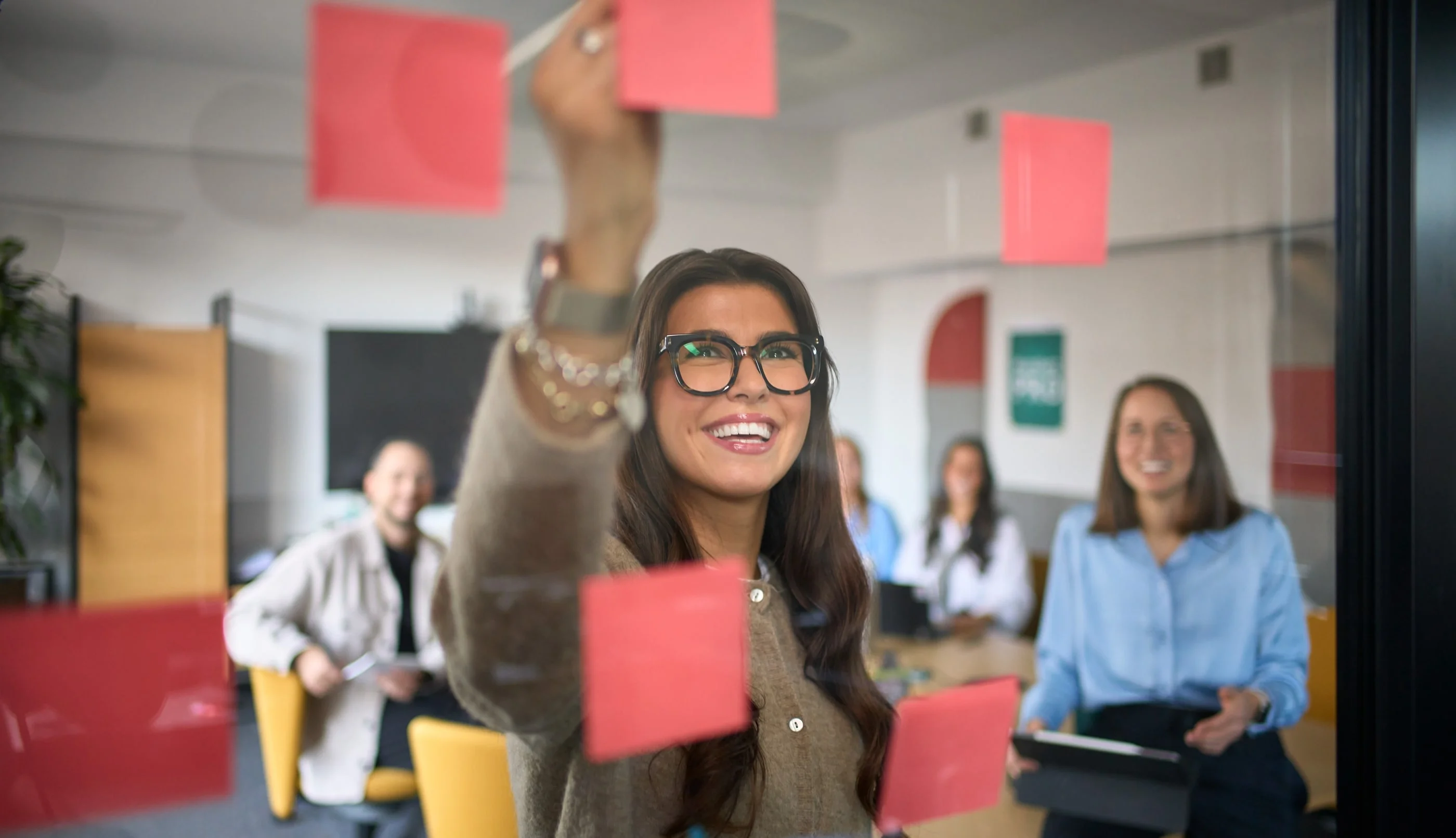 Eine lächelnde Frau mit Brille befestigt bunte Haftnotizen an einer Glasscheibe in einem Besprechungsraum. Im Hintergrund sitzen mehrere Personen und beobachten sie, während sie an einem Projekt arbeiten. Die Atmosphäre ist kreativ und kollaborativ.