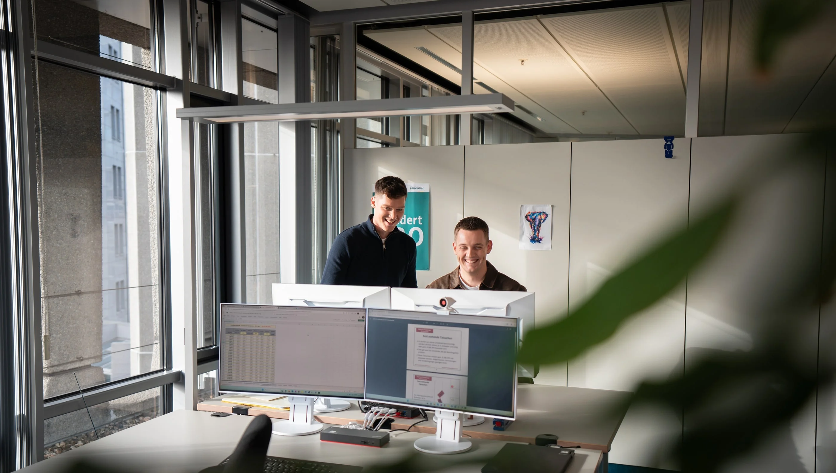 Zwei Männer stehen vor einem Computerarbeitsplatz in einem modernen Büro. Sie schauen auf den Bildschirm, während sie miteinander sprechen. Der Raum hat große Fenster, die natürliches Licht hereinlassen. Im Hintergrund sind Büromöbel und eine Pflanze sichtbar.