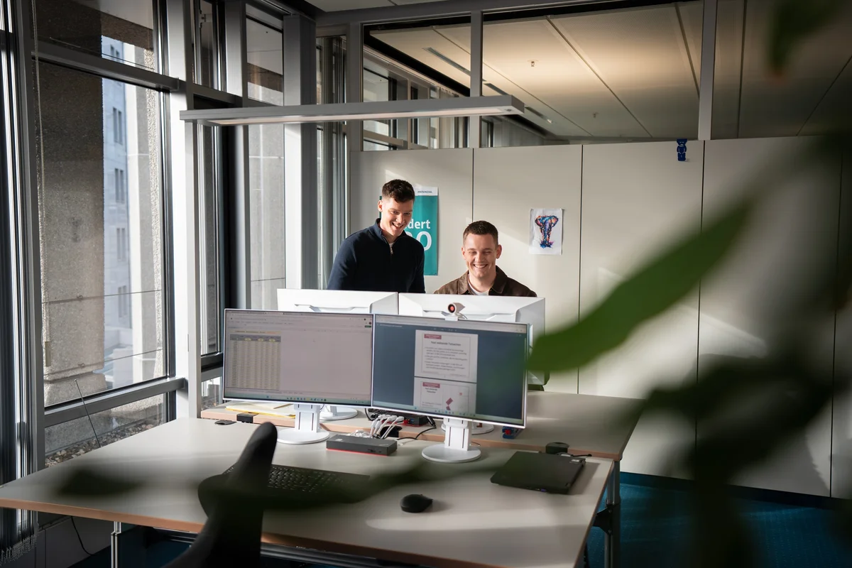Zwei Männer stehen vor einem Computerarbeitsplatz in einem modernen Büro. Sie schauen auf den Bildschirm, während sie miteinander sprechen. Der Raum hat große Fenster, die natürliches Licht hereinlassen. Im Hintergrund sind Büromöbel und eine Pflanze sichtbar.
