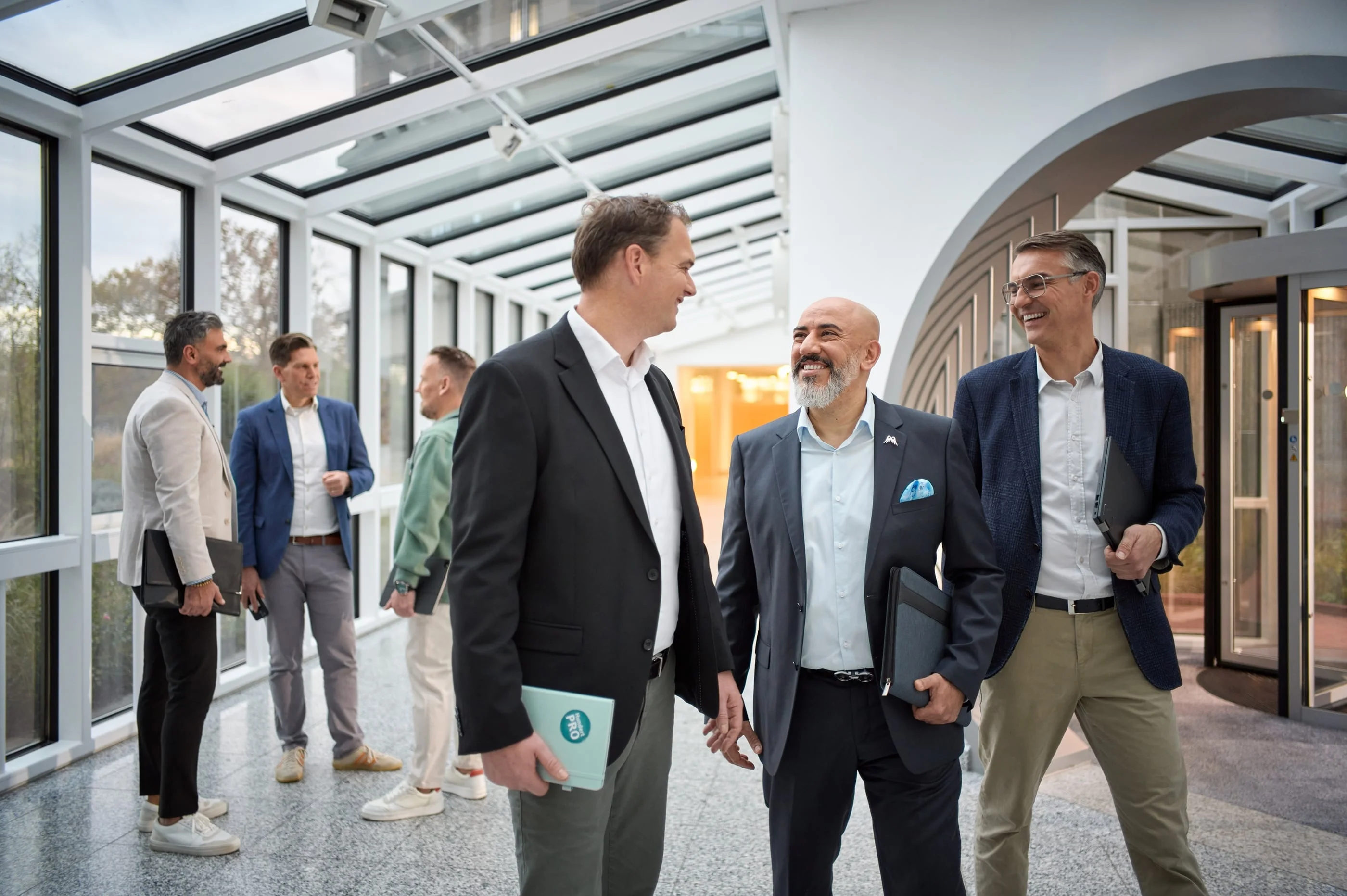 Eine Gruppe von sechs Personen steht in einem modernen, hellen Flur mit Glaswänden. Drei Männer im Vordergrund unterhalten sich freundlich, während die anderen im Hintergrund stehen und ebenfalls in einem Gespräche vertieft sind. Alle tragen Businesskleidung.