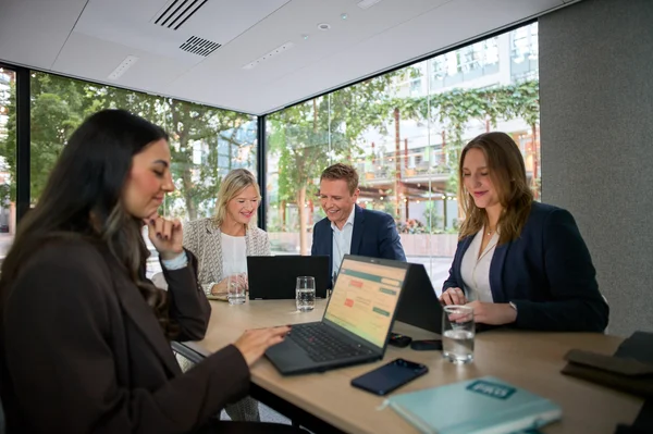 Eine Gruppe von vier Personen sitzt an einem Tisch in einem Konferenzraum. Sie arbeiten an Laptops und tauschen Ideen aus. Die Atmosphäre ist freundlich und konzentriert, mit Pflanzen und großen Fenstern im Hintergrund, die auf einen Außenbereich blicken.