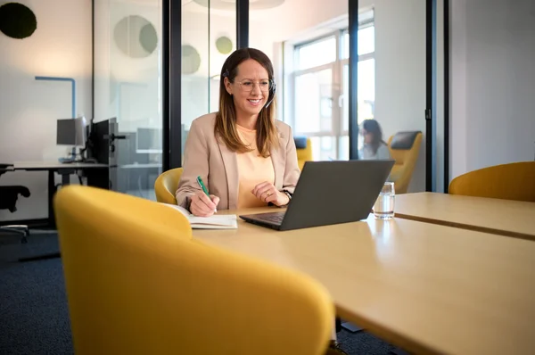 Eine lächelnde Frau sitzt an einem Tisch in einem modernen Büro und arbeitet an ihrem Laptop. Sie hat Kopfhörer auf und hält einen Stift in der Hand, während sie Notizen macht. Im Hintergrund sind verschwommene Figuren zu sehen.