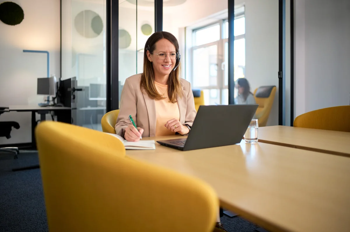 Eine lächelnde Frau sitzt an einem Tisch in einem modernen Büro und arbeitet an ihrem Laptop. Sie hat Kopfhörer auf und hält einen Stift in der Hand, während sie Notizen macht. Im Hintergrund sind verschwommene Figuren zu sehen.