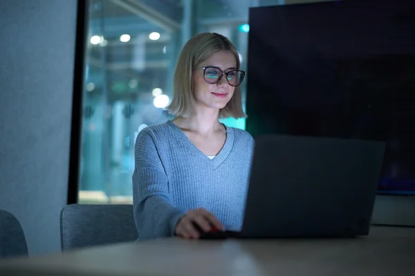 Eine junge Frau mit blonden Haaren und Brille sitzt an einem Tisch und arbeitet konzentriert an einem Laptop. Sie trägt einen hellblauen Pullover.