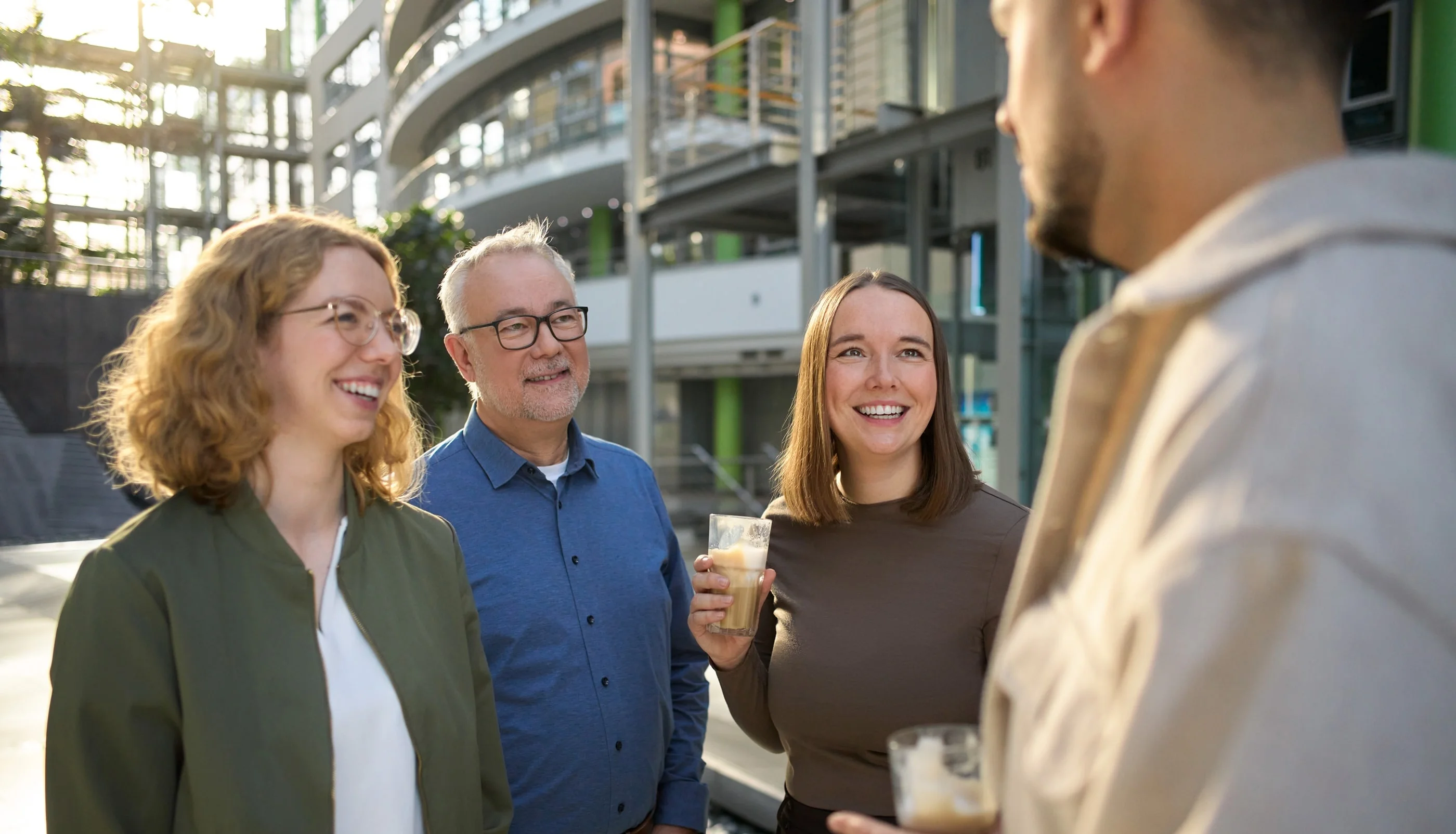 Vier Personen unterhalten sich lächelnd im Freien. Zwei Frauen und ein Mann stehen zusammen, während eine weitere Person, die ihnen gegenübersteht, spricht. Alle halten Getränke in der Hand. Die Umgebung wirkt modern und hell.