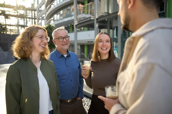 Vier Personen unterhalten sich lächelnd im Freien. Zwei Frauen und ein Mann stehen zusammen, während eine weitere Person, die ihnen gegenübersteht, spricht. Alle halten Getränke in der Hand. Die Umgebung wirkt modern und hell.