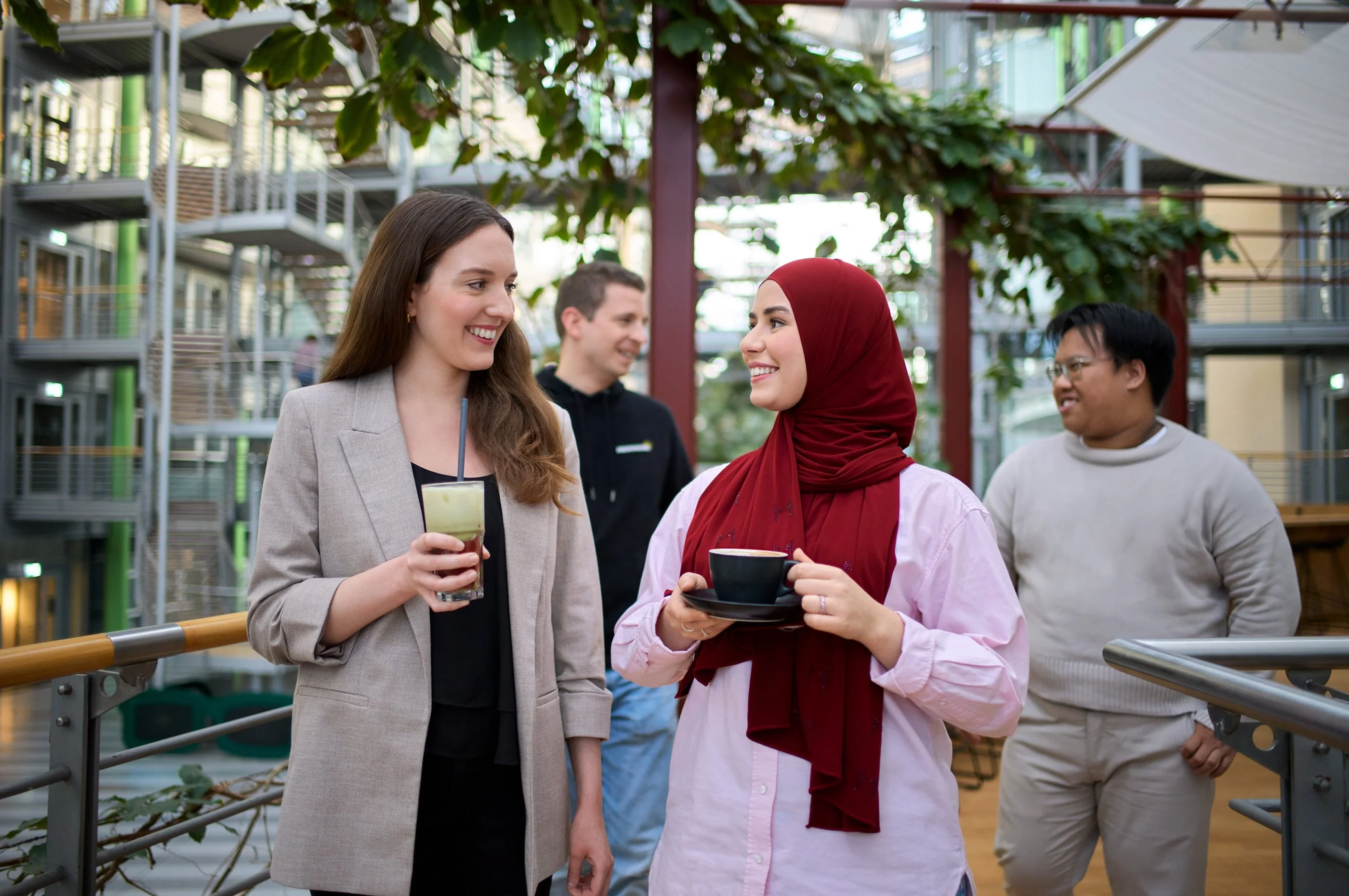 Zwei Frauen unterhalten sich auf einer überdachten Terrasse, während sie Getränke halten. Die eine trägt einen Blazer und die andere ein rotes Kopftuch. Im Hintergrund sind zwei Männer zu sehen, die ebenfalls an der Unterhaltung teilnehmen. Die Atmosphäre wirkt freundlich und entspannt.