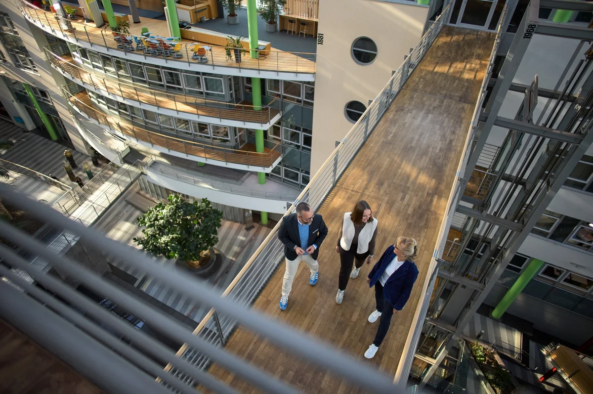 Drei Personen stehen auf einer Brücke in einem modernen Gebäude, umgeben von großen Fenstern und Pflanzen. Sie scheinen ein Gespräch zu führen. Die Architektur ist offen und hell, mit mehreren Etagen sichtbar.