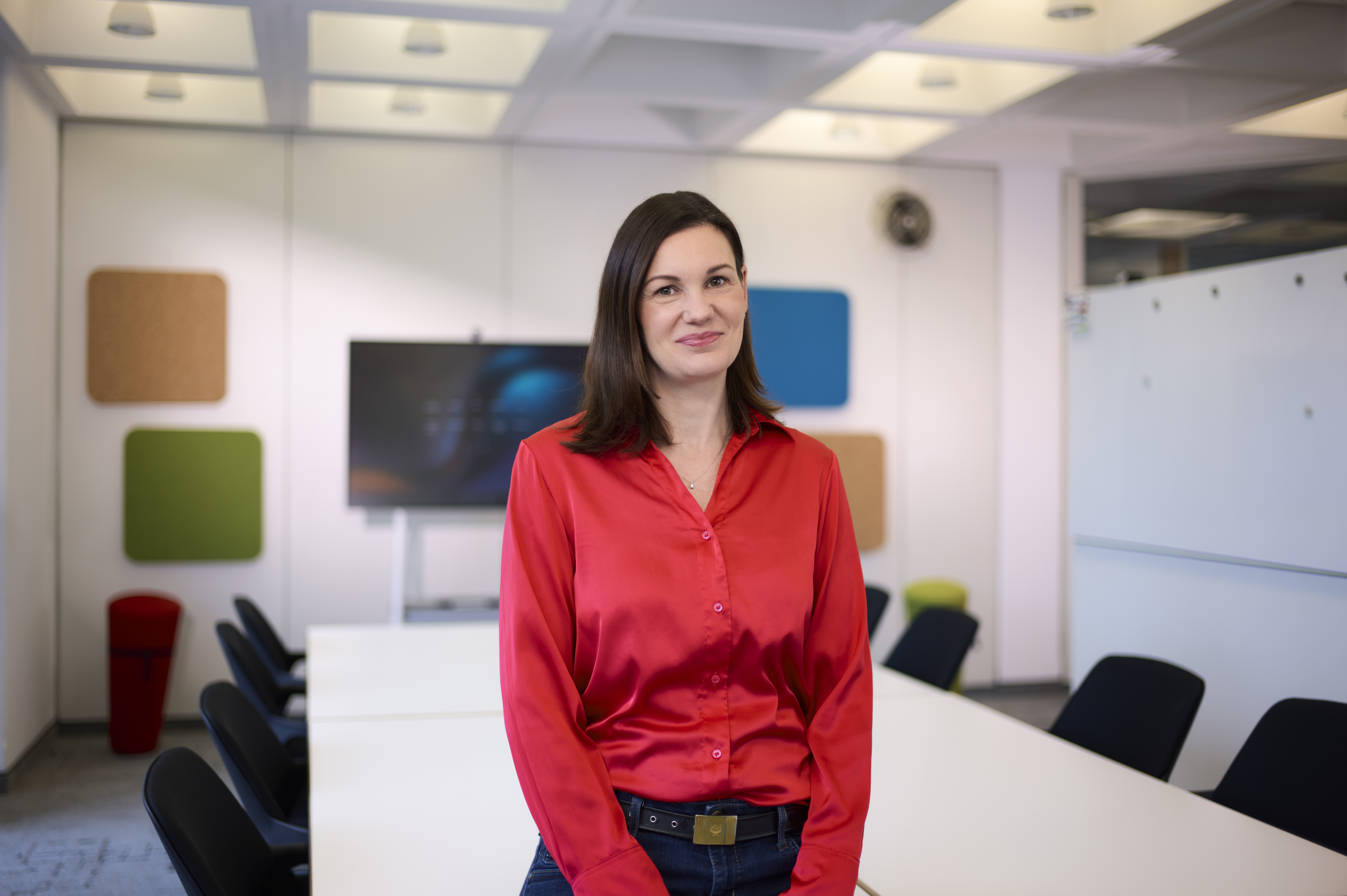 Eine Frau in einer roten Bluse steht in einem Konferenzraum mit einem langen Tisch und mehreren Stühlen. Im Hintergrund ist ein Bildschirm sowie farbige Wandpaneele zu sehen. Das Licht im Raum ist hell und gleichmäßig.