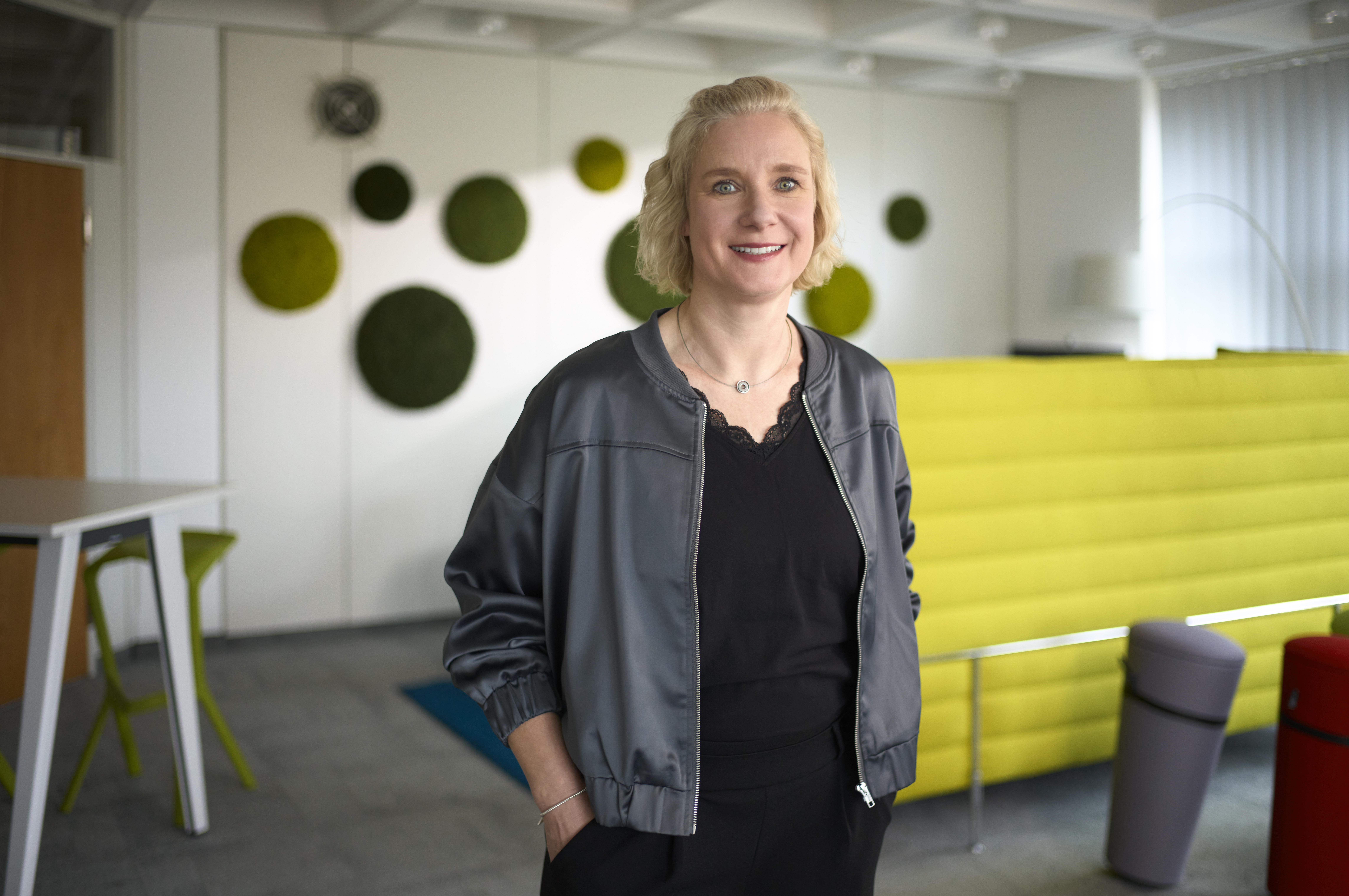 Eine lächelnde Frau steht in einem modernen Büro mit grünen Deko-Elementen an der Wand und einem gelben Sofa im Hintergrund.