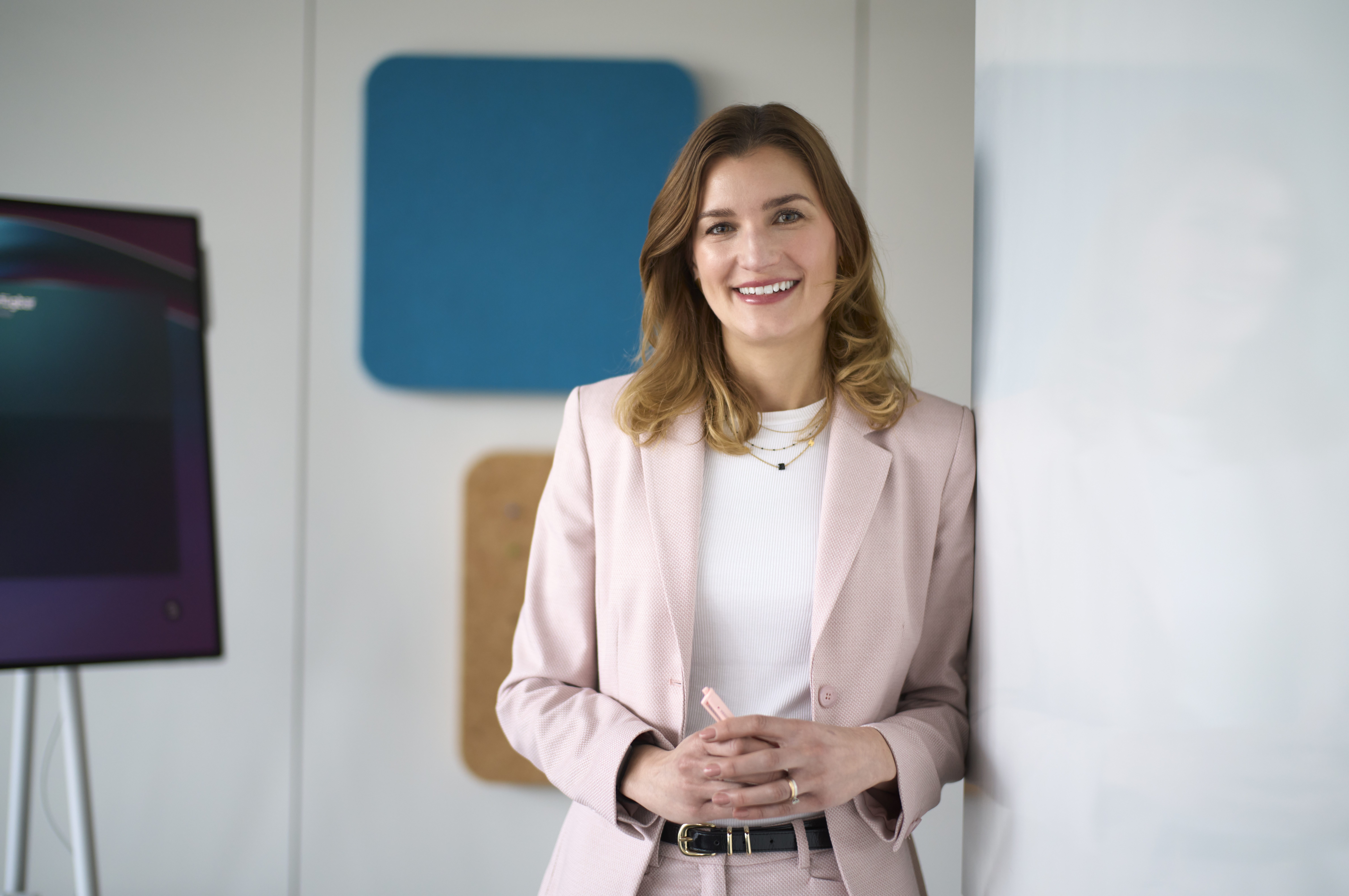 Eine lächelnde Frau in einem hellrosa Anzug steht in einem modernen Büro. Sie hält ihre Hände gefaltet vor sich. Im Hintergrund sind eine Leinwand und bunte Paneele an der Wand zu sehen.