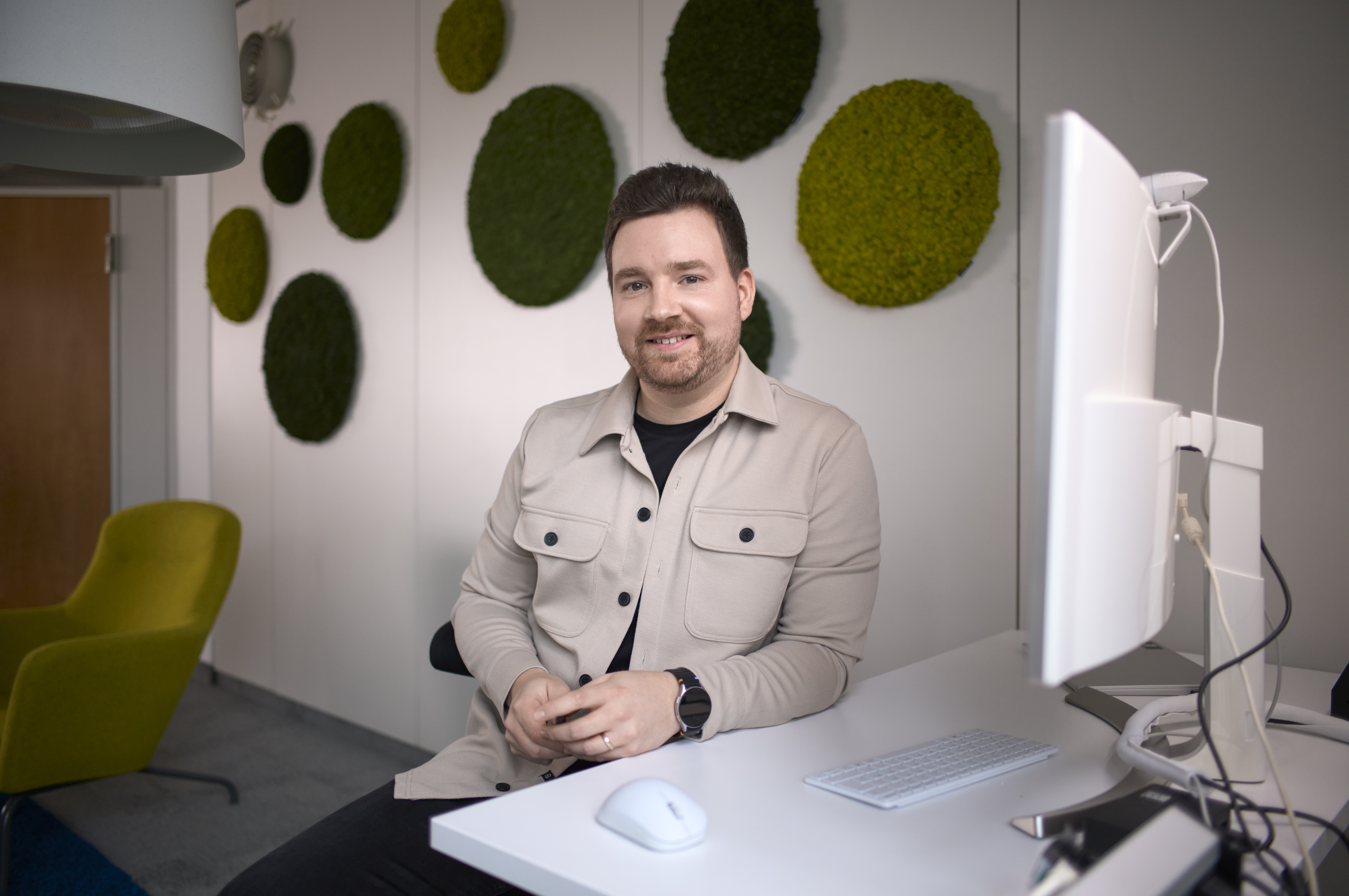 Ein Mann sitzt lächelnd an einem Schreibtisch mit einem Computer. Im Hintergrund sind runde grüne Moosdekorationen an der Wand und ein gelber Stuhl zu sehen. Er trägt eine beige Jacke und eine Uhr.