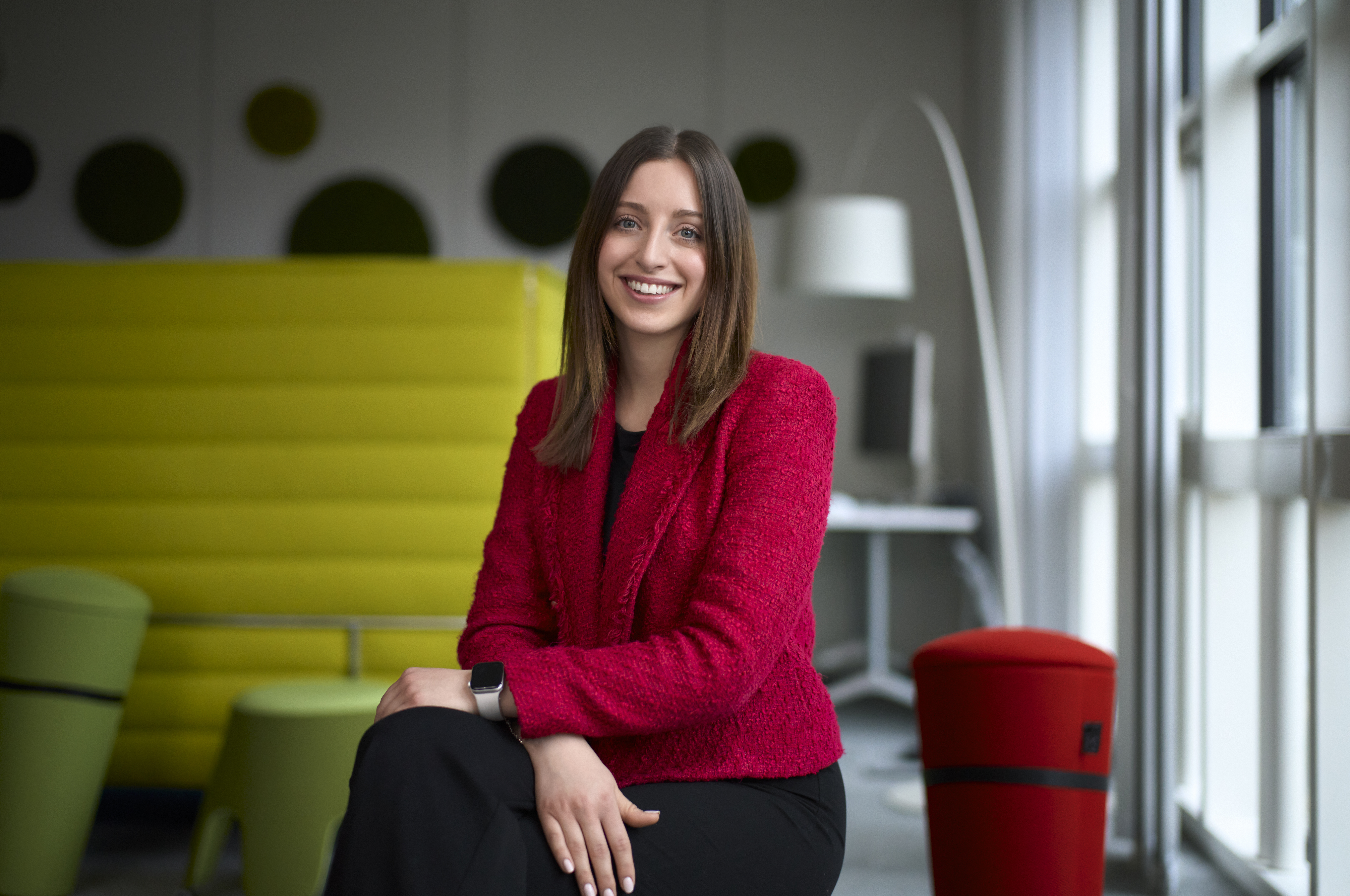 Frau in rotem Blazer sitzt lächelnd in einem modernen Büro mit farbigen Möbeln im Hintergrund. Sie trägt eine Smartwatch.