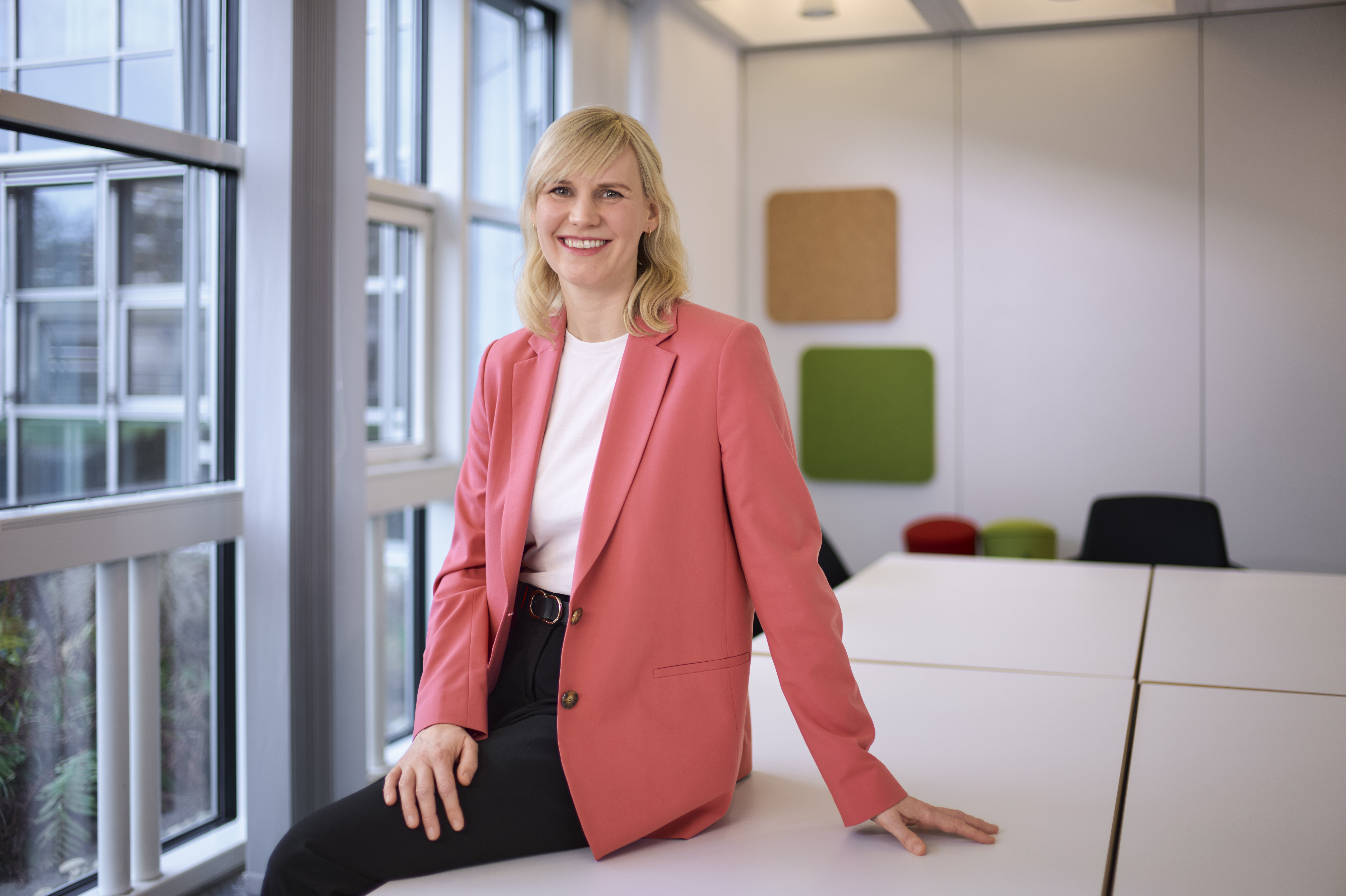 Eine Frau mit blonden Haaren sitzt auf einem Tisch in einem modernen Raum. Sie trägt einen pinken Blazer, ein weißes T-Shirt und schwarze Hosen. Im Hintergrund sind Fenster und bunte Wanddekorationen zu sehen. Sie lächelt in die Kamera.