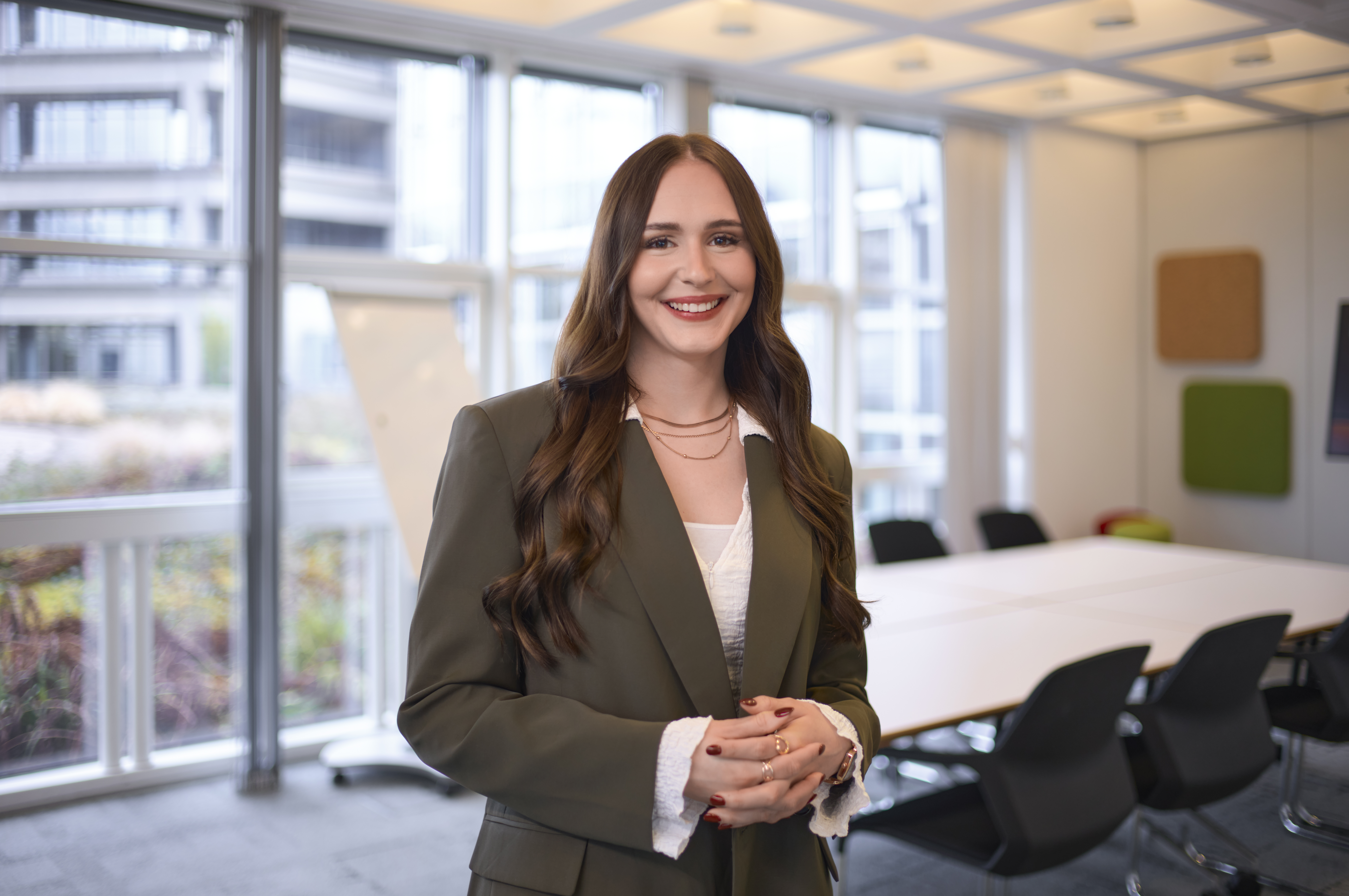 Eine Frau mit langen braunen Haaren und einem grünen Blazer lächelt in einem modernen Büro mit großen Fenstern und einem Konferenztisch im Hintergrund.