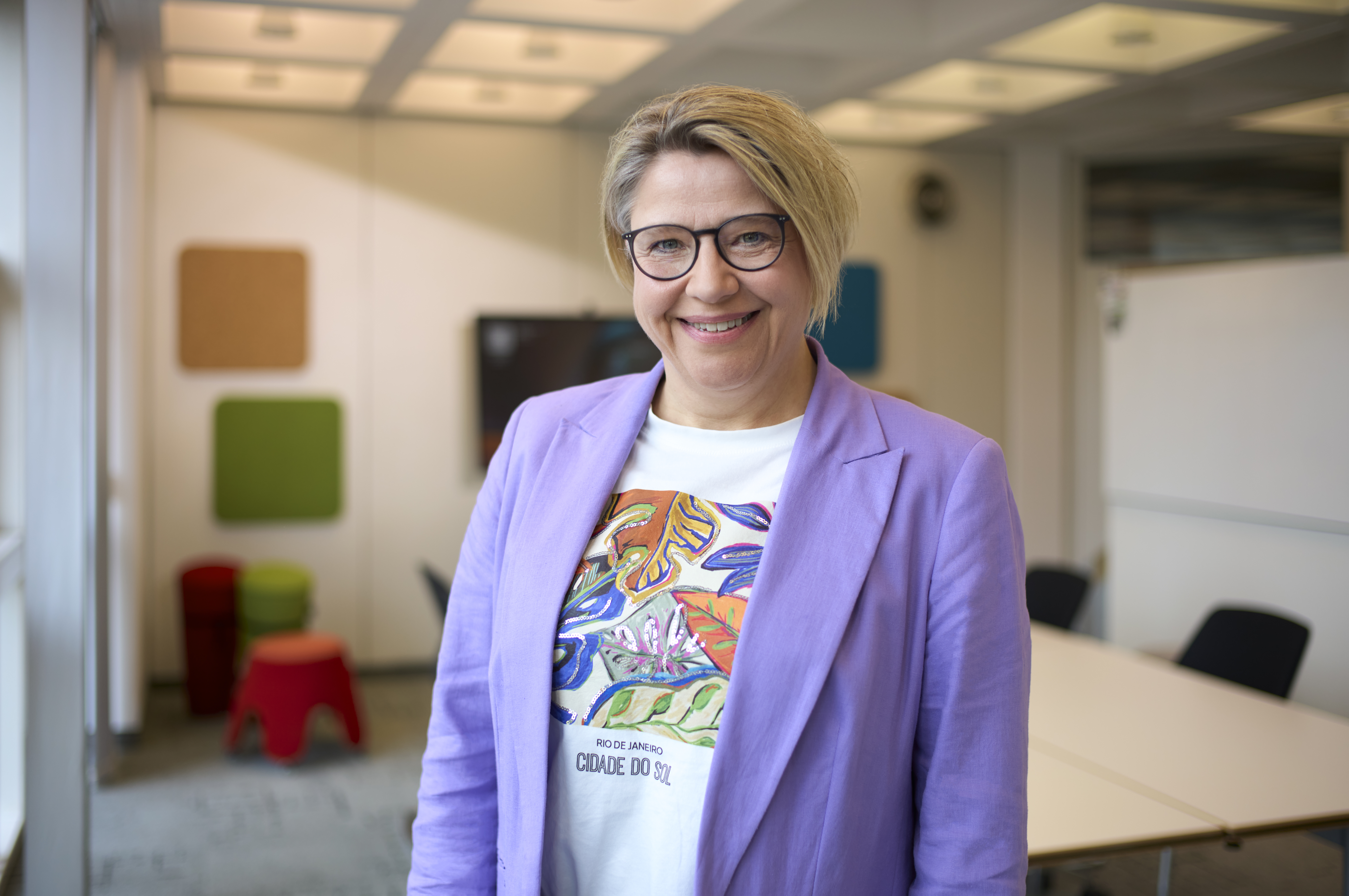 Frau in einem lila Blazer steht in einem modernen Büro, lächelt und schaut in die Kamera. Sie trägt ein buntes T-Shirt mit tropischem Design. Im Hintergrund sind bunte Wandtafeln und Büromöbel sichtbar.